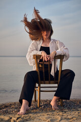 Young woman sits on chair facing calm sea, symbolizing solitude and introspection. Her relaxed...