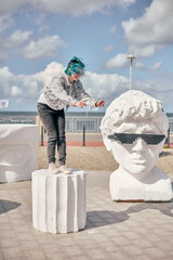 Young male with blue hair jumps over large white head sculpture in sunny promenade. Concrete...