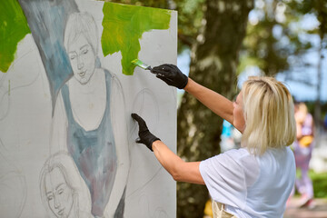 Young adult female painting on outdoor canvas. Blonde hair, white shirt, gloves. Background...