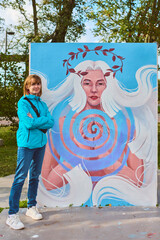 Young female with light brown hair stands beside vibrant mural featuring mystical figure. Scene...