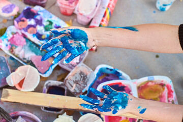 Adult hands covered in vivid blue paint, with scattered art supplies outdoors. Soft lighting casts...