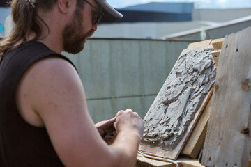 Adult male artist sculpting clay relief outdoors. Strong sunlight, textured board background, close-up shot, focused on intricate details