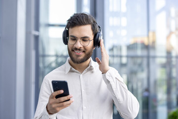 A man wearing headphones listens to music on his phone, smiling near a window.