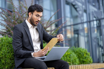 An astonished businessman sits outside with a laptop, examining an envelope with a look of...