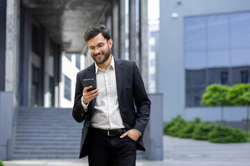 A smiling businessman in a suit looks at his phone outdoors in front of a modern building, a...