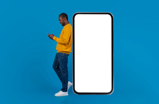 Happy stylish middle-aged african american man leaning on big smartphone with white empty screen and using cell phone, using newest dating mobile app, blue background, copy space, full length, mockup