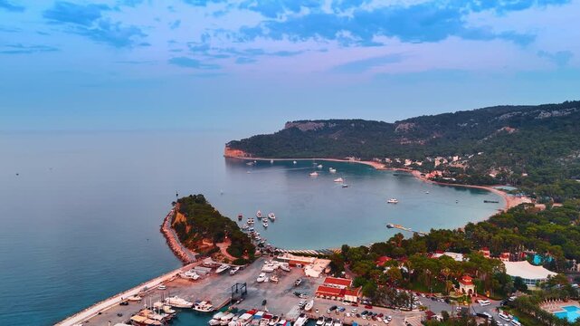 Multiple yachts stand at the berths and on the waterscape. Picturesque rocky shore of the the Mediterranean coast of Turkey at dusk time. Aerial view.