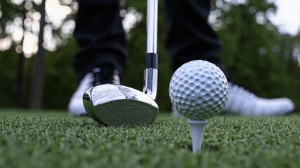 Low angle view of a golfer's feet and lower legs, positioned behind a golf ball on a tee, with the club head poised to strike, ready for the swing on a meticulously maintained golf course