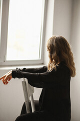 Rare view of young woman with wawy hair sitting on the chair near window and looking in distance, planning common future, enjoying peaceful moment. Space for text