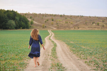 Rear view of a beautiful blonde woman walking on winding dirt road on nature background. Woman in polka-dot dress walking outdoor. Horizontal photo, space for text