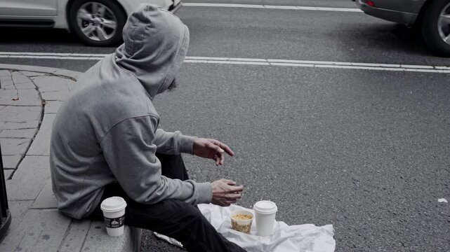 Homeless man sits on the curb of a busy city street, eating a meager meal while cars pass by, highlighting the struggles of urban poverty and social disparity