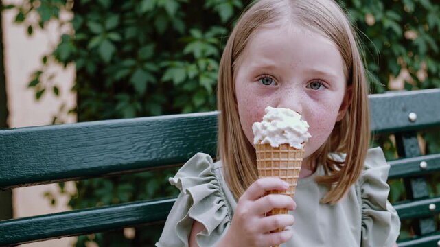 Freckled young girl sits on a park bench, indulging in a vanilla ice cream cone, her eyes closed in pure bliss as she savors each bite against a backdrop of green foliage. happy childhood