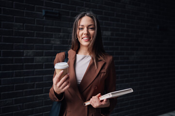 Smiling businesswoman holding coffee and books by brick wall