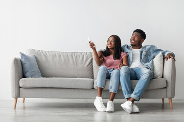 Split System. Satisfied African American woman holding remote control relaxing under the air conditioner, happy millennial black couple sitting on couch in living room, free copy space