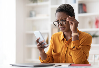 Confused pretty millennial black woman in casual outfit looking at cell phone screen and touching...