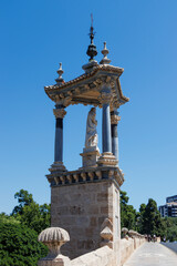 A detailed view of a historical sculpture on a stone tower in Valencia, adorned with classical architecture, against a clear blue sky, highlighting cultural heritage.