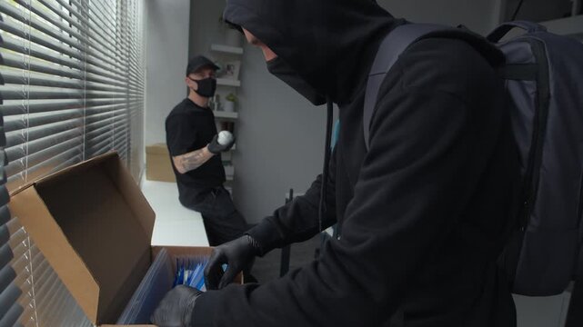 Side view of masked male criminal wearing gloves rummaging through folders with documents in box searching for confidential data while discussing crime plan with laid back accomplice in office
