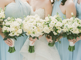 Bridesmaids in light blue dresses hold white floral bouquets at a wedding ceremony in a garden...