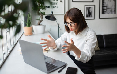 Angry frowning young businesswoman working online on laptop pc computer, getting video call or reading emails and gesturing, sitting at coworking space, copy space