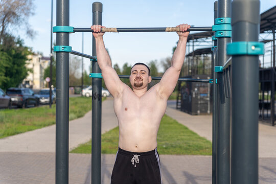 Muscular man doing pull-ups on outdoor gym equipment. Shirtless, he showcases defined upper body strength. Set in a sunny park, the photo captures a healthy lifestyle