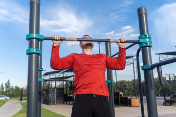 Man in red shirt doing pull-ups at an outdoor gym. The blue sky suggests a sunny day for a workout and active lifestyle