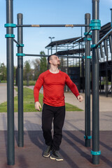Man in red shirt doing pull-ups at an outdoor gym. The blue sky suggests a sunny day for a workout and active lifestyle