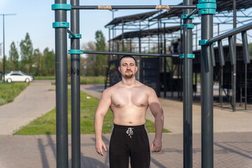 Muscular man doing pull-ups on outdoor gym equipment. Shirtless, he showcases defined upper body strength. Set in a sunny park, the photo captures a healthy lifestyle