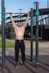 Muscular man doing pull-ups on outdoor gym equipment. Shirtless, he showcases defined upper body strength. Set in a sunny park, the photo captures a healthy lifestyle