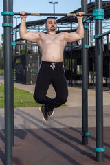 Muscular man doing pull-ups on outdoor gym equipment. Shirtless, he showcases defined upper body strength. Set in a sunny park, the photo captures a healthy lifestyle