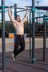 Muscular man doing pull-ups on outdoor gym equipment. Shirtless, he showcases defined upper body strength. Set in a sunny park, the photo captures a healthy lifestyle