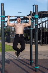 Muscular man doing pull-ups on outdoor gym equipment. Shirtless, he showcases defined upper body strength. Set in a sunny park, the photo captures a healthy lifestyle