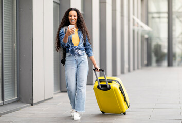 Fototapeta na wymiar Cheerful pretty curly middle eastern young woman in comfy casual outfit going vacation alone, walking by airport with yellow luggage, using cell phone, online check-in, copy space, full length