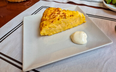A slice of Spanish tortilla served on a white plate with a dollop of creamy sauce. The dish...