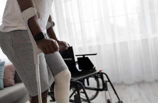 Trauma, post-incident fracture of bone, exercise and recovery at home. Tense millennial african american man with broken leg in cast walks with crutches to wheelchair in living room interior, cropped - Powered by Adobe