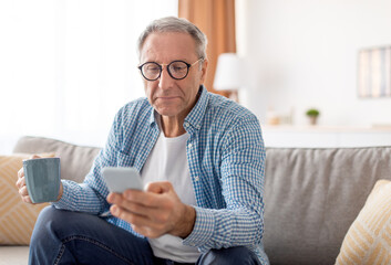Portrait Of Mature Guy In Eyeglasses Using Mobile Phone With New Application Sitting On Couch, Reading News Or Message Drinking Coffee. Gadgets And Mobile Communication, Adult People And Tech Concept