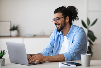 Smiling western freelancer guy working on laptop at home office, sitting at desk using computer,...