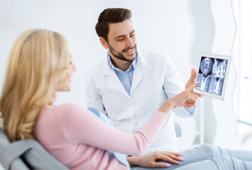 Male dentist showing female patient jaw x-ray on digital tablet, dental clinic interior. Bearded man stomatologist having conversation with lady in dental chair, pointing at gadget, side view