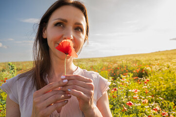 Serene woman with eyes closed enjoying the fragrance of a red poppy flower in a sunlit meadow....