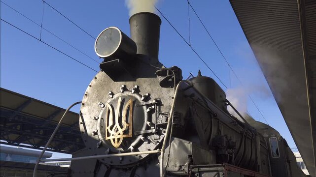 Close-up of an old black steam locomotive with a golden Ukrainian trident on the front under a blue sky. Fixed camera