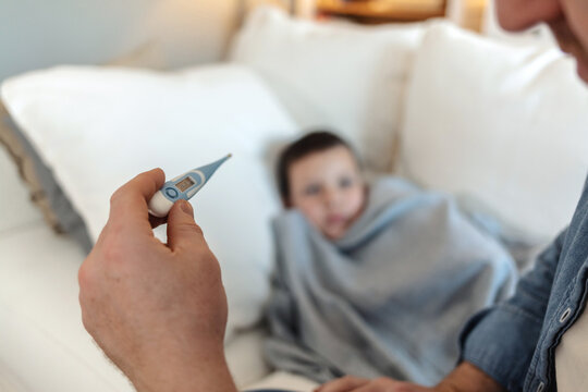 Young father checking temperature of sick son lying while lying in bed. Father checking her sick sons temperature.