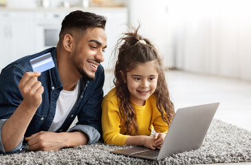 Portrait of happy arab dad and cute little daughter with laptop and credit card making online shopping together at home, middle eastern family relaxing on carpet with computer, closeup shot