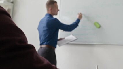Rack focus shot of male manager writing on whiteboard discussing with team weekly work plan while colleague taking notes during meeting in conference room at office