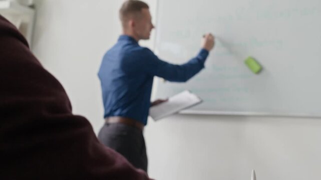 Rack focus shot of male manager writing on whiteboard discussing with team weekly work plan while colleague taking notes during meeting in conference room at office