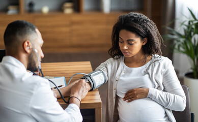 African american man doctor gynecologist checking young pregnant woman heart rate and blood pressure, using tonometer, modern clinic interior, copy space. Medical care during pregnancy