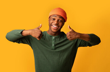 Naklejka premium All Is Great. Cheerful Black Millennial Guy Showing Thumbs Up At Camera, Positive African American Hipster Man Gesturing Sign Of Approval, Recommending Something While Standing Over Yellow Background
