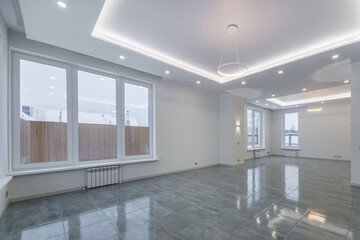 A spacious living room in a country house without furniture. Grey tiled floor, white walls, spotlights. Panoramic windows.