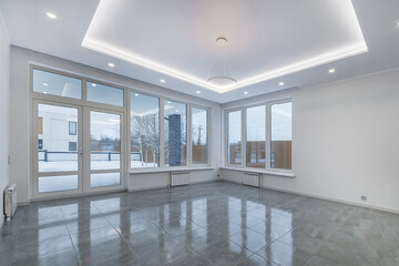 An empty spacious bright room with panoramic windows. Grey floor tiles, white decor, spot ceiling lights.