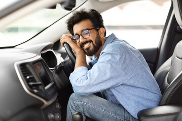 Automobile renting, buying, leasing. Thrilled young indian guy hugging steering wheel of his brand...