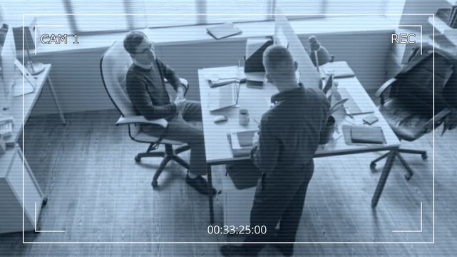 Security camera footage of male office worker chatting with colleague discussing job tasks while sitting at desk with laptop in modern workspace