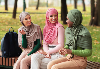 Three Diverse Muslim Ladies In Hijab Talking Enjoying Conversation And Autumn Day Sitting On Bench In City Park. Modern Islamic Female Students Spending Time Chatting Outdoors
